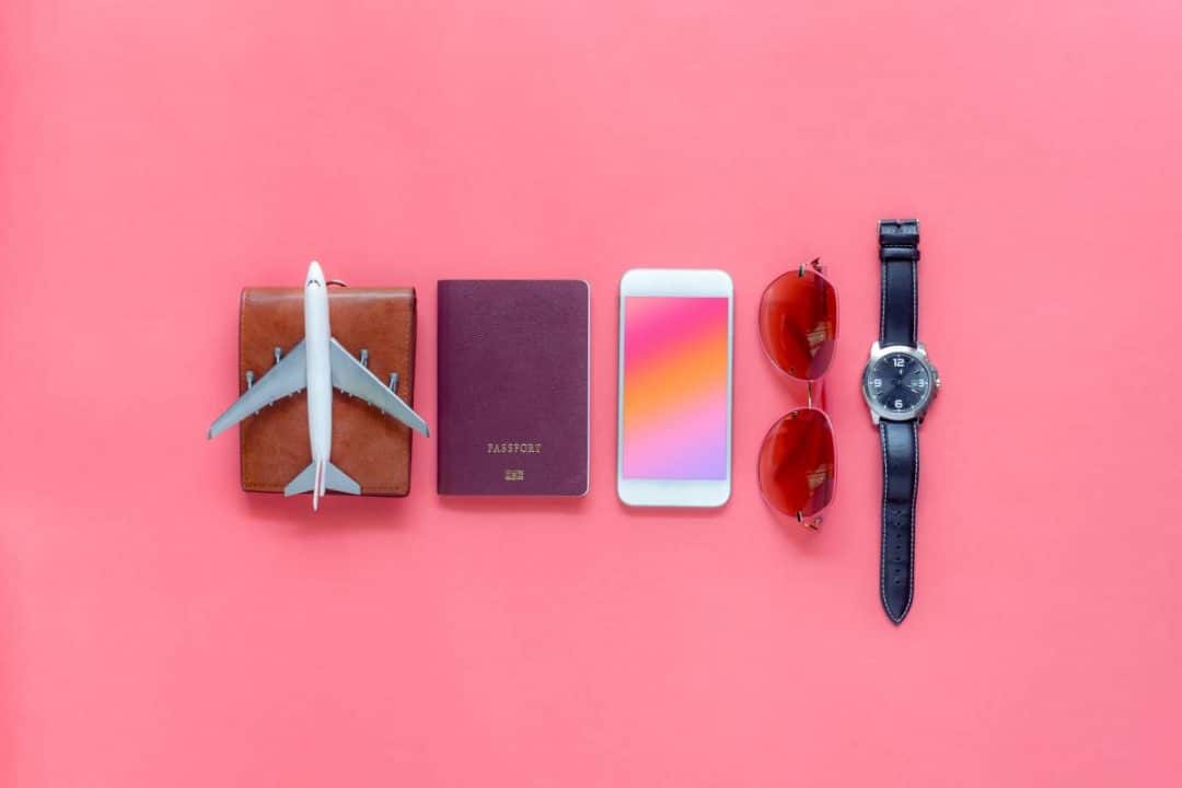 Pink background with small items including a toy airplane on a wallet, passport, iPhone, sunglasses and a watch