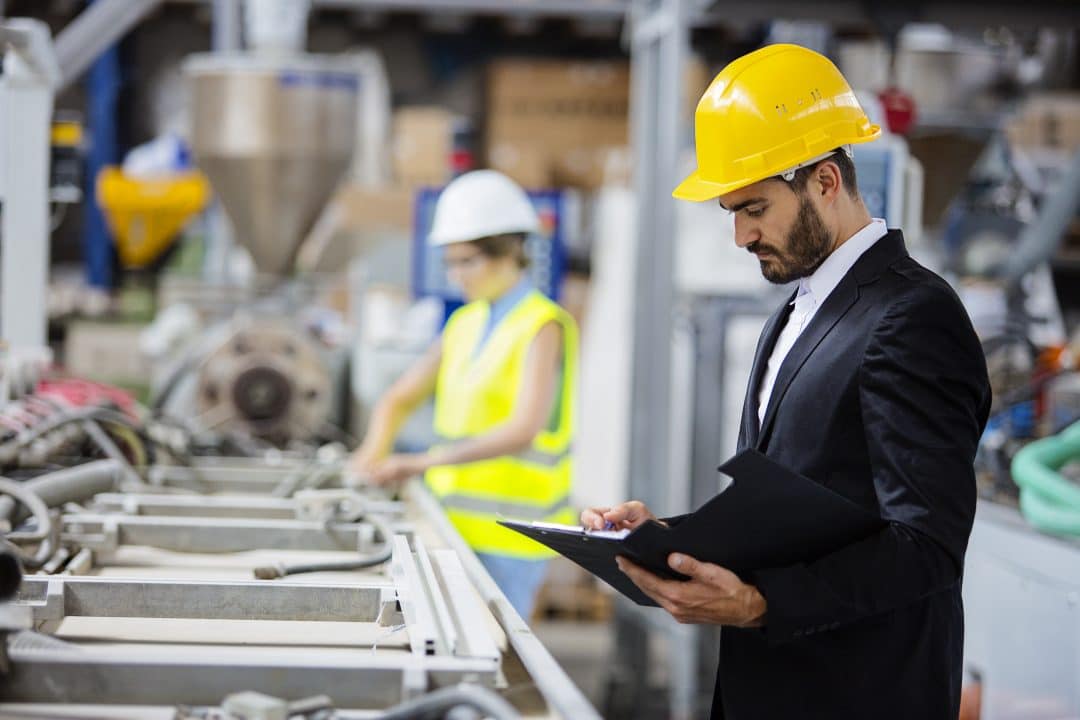 Man in a suit and yellow hard hat looking at parts in a factory and taking notes