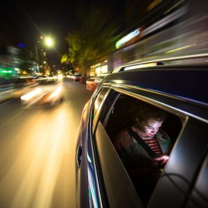 A woman sits in the back of a car, possibly a rideshare, looking at her mobile phone. The lights of New York City streak past.