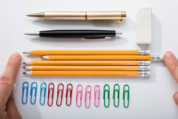 Lined up pens, pencils and paper clips. One of the pencils is out of line. A person is pushing it in line.
