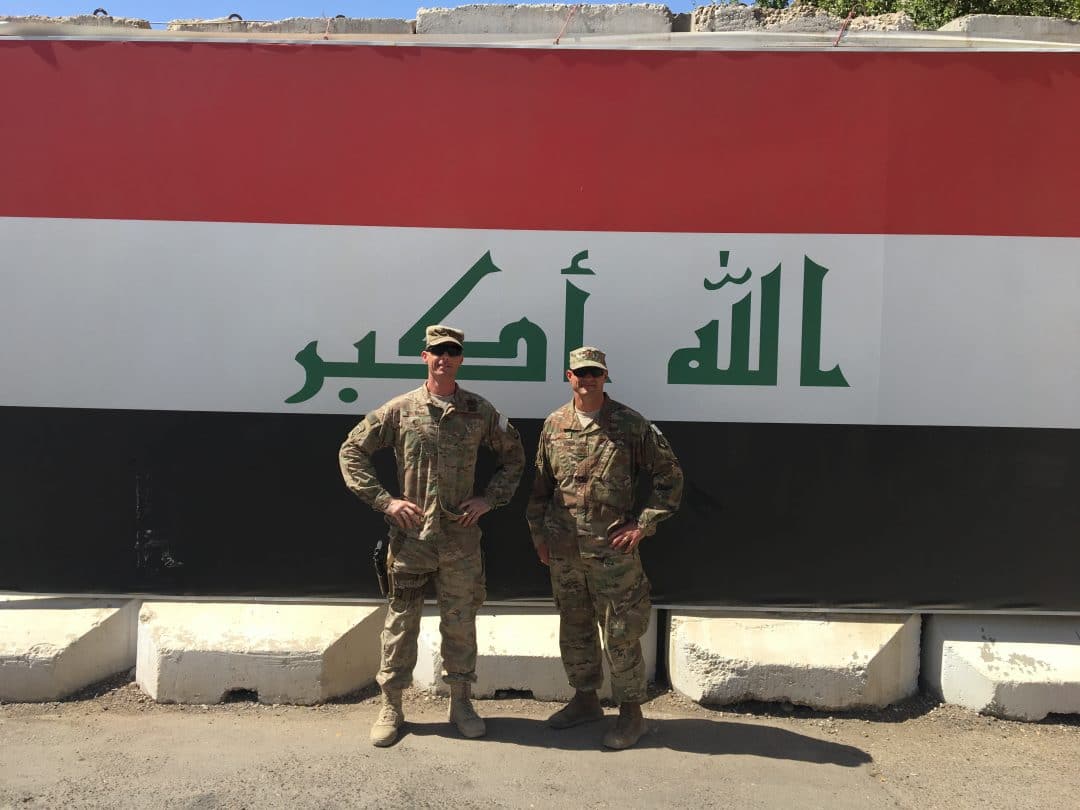Two men in uniform standing in front of a mural of the Iraqi flag in Baghdad.