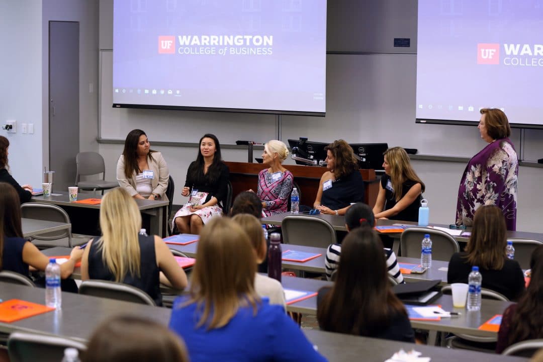 Six women at the front of a classroom speaking to a room of female students