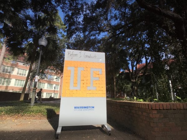 A large white board on wheels sits in a green courtyard. The white board reads Before I graduate with the UF logo, which is made of post it notes