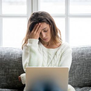 Young woman sitting on a couch looking at a computer. She holds her hand up to her forehead looking stressed.