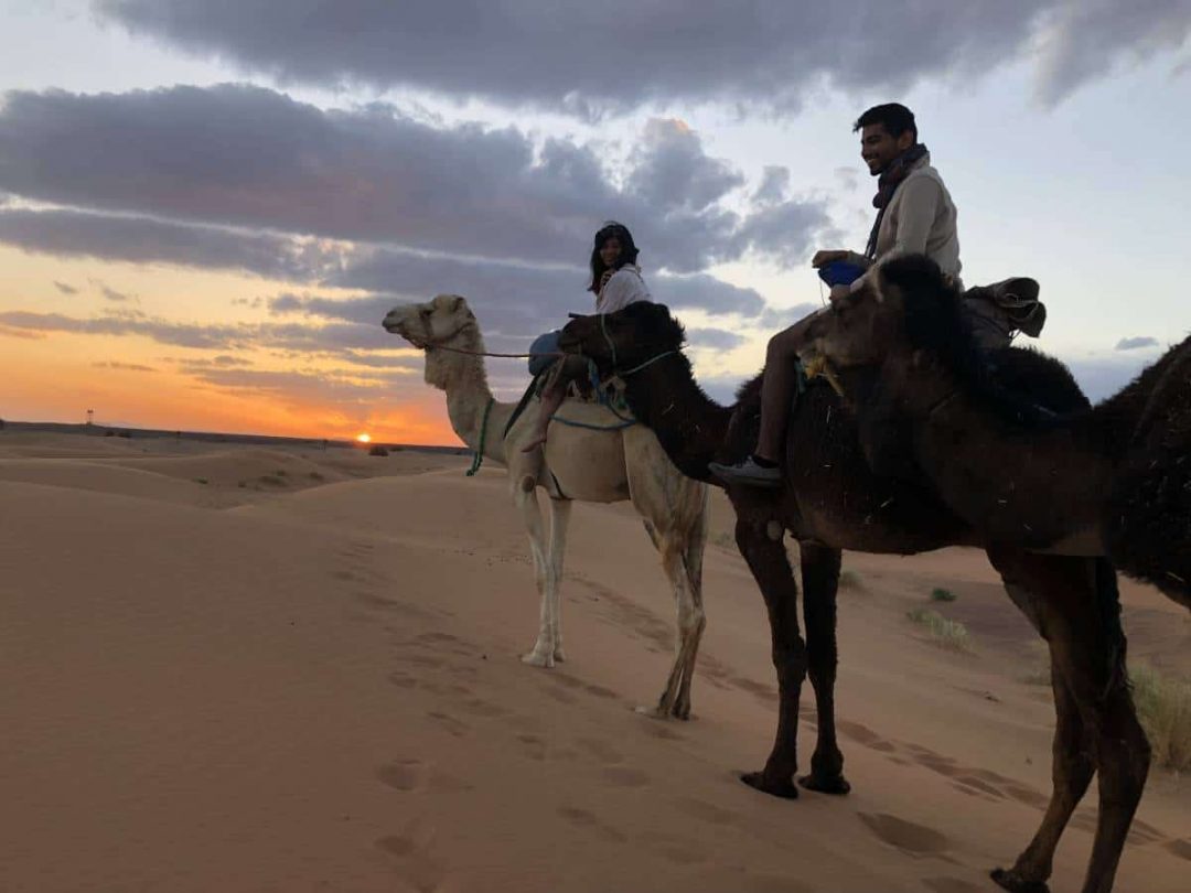 Two students riding camels on sandy dunes at sunset