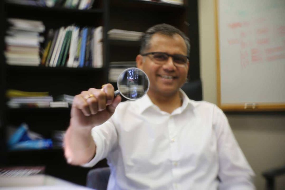 Warrington faculty member Anuj Kumar sitting in his office by a bookshelf holding up a magnifying class that reflects the courtyard outside of his office.