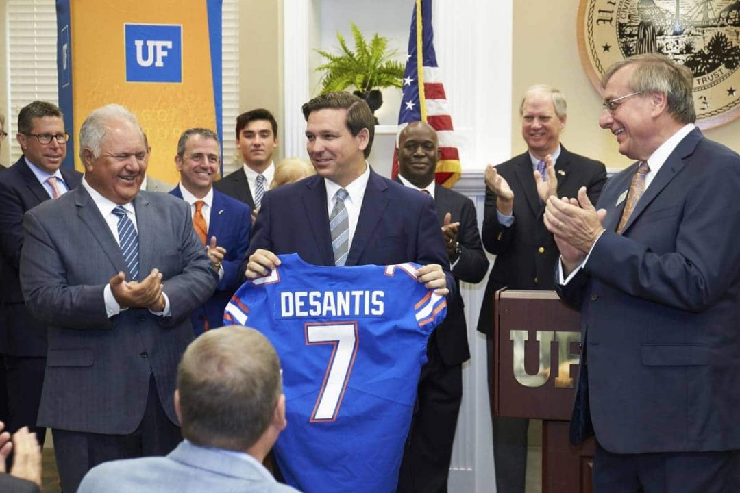 Florida Governor Ron DeSantis holds up a UF football jersey with his last name and the number 7. He is flanked by More Hossini and UF President Kent Fuchs who are clapping