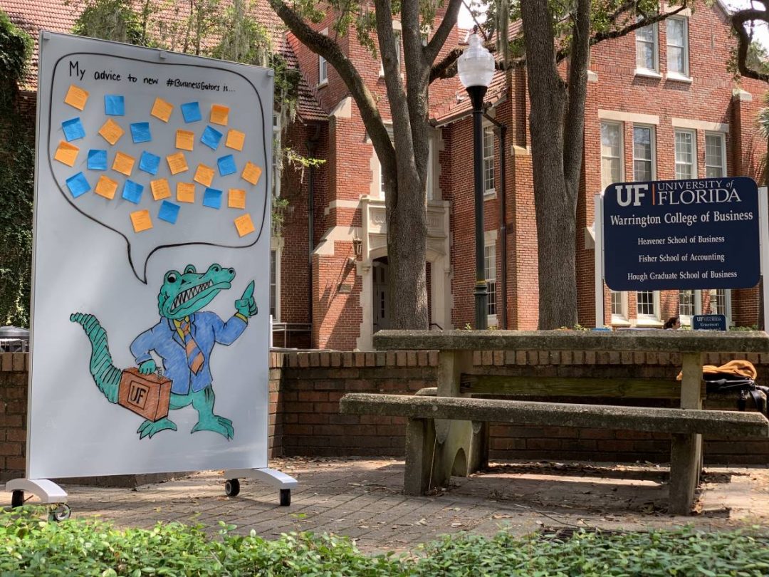 Whiteboard with drawing of a Gator with a speech bubble above his head with advice. The whiteboard is outside in a courtyard next to a bench in front of a sign that says Warrington College of Business