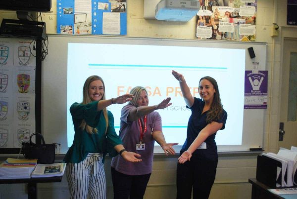 Three women stand in front of a projection screen doing the Gator Chomp
