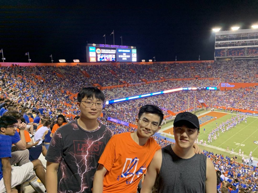 Three Asian students stand up for a photo during a football game in a crowded stadium.