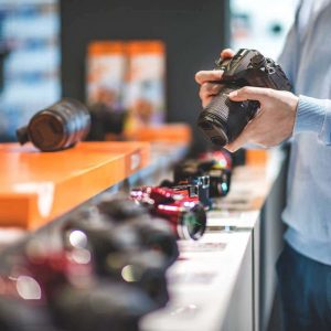 Unrecognizable male customer is holding black DSLR camera in the store. Row of other camera models is visible on the shelf.