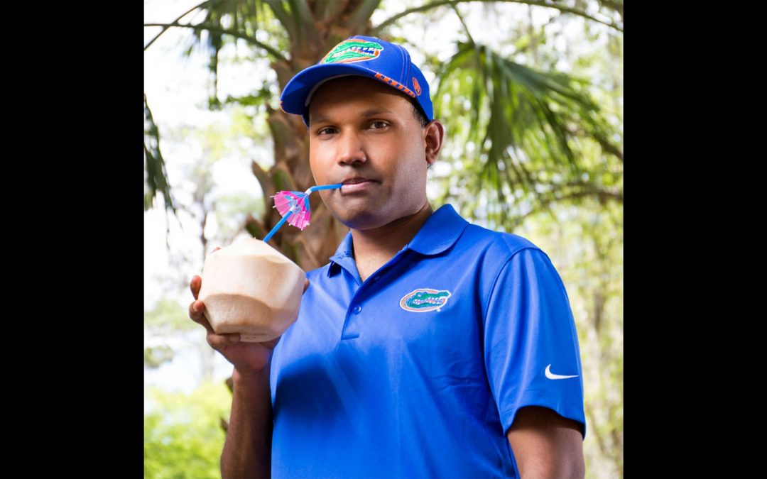 Cocovana founder Sheldon Barrett, wearing a blue Gator shirt and hat, drinks out of a coconut through a straw with an umbrella.