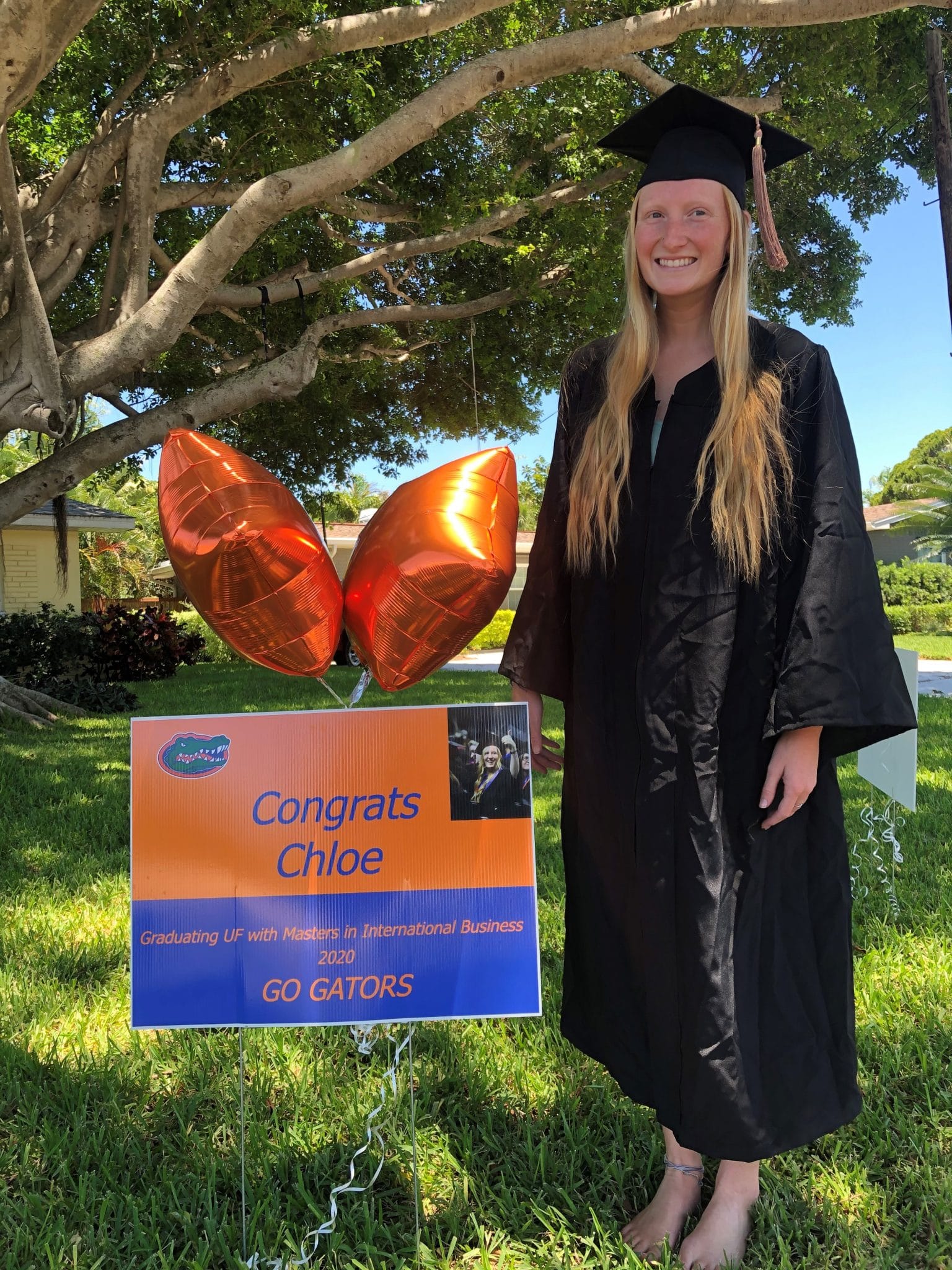Chloe Beaver stands in her cap and gown outside next to an orange and blue sign that reads Congratulations Chloe