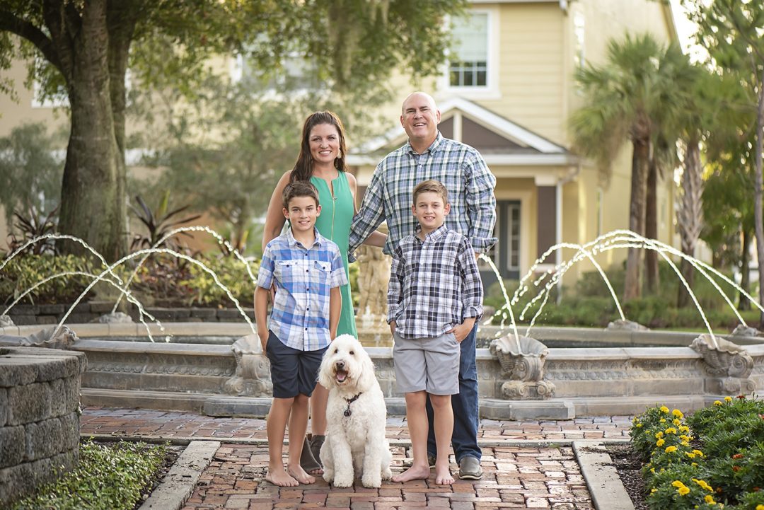 Cindy Trautmann poses with her husband, two sons and dog in front of a fountain, garden and yellow house