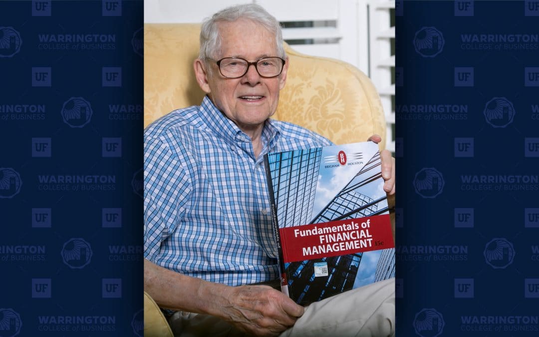 Eugene Brigham sits in a chair holding a copy of his textbook Fundamentals of Financial Management