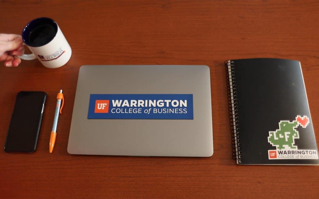 Birds eye view of a lap top with a Warrington sticker, notebook, pen, cell phone and coffee mug with a hand on the handle.