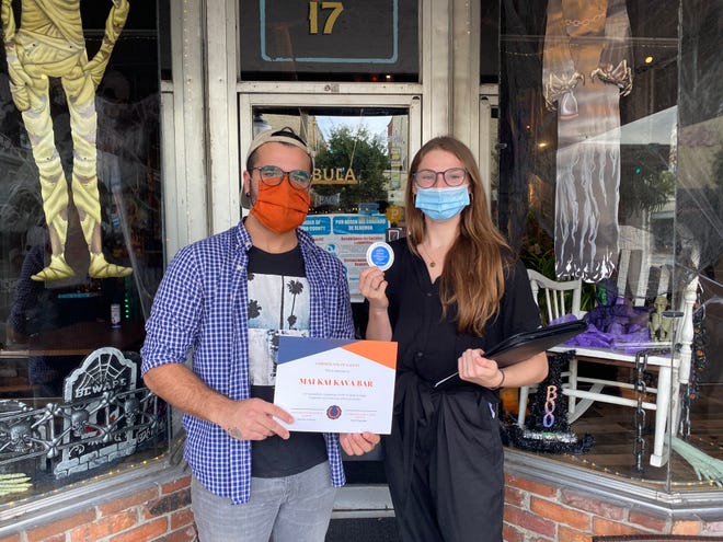 Two people stand in a doorway. Both wear masks as one holds up a certificate and the other holds a sticker.