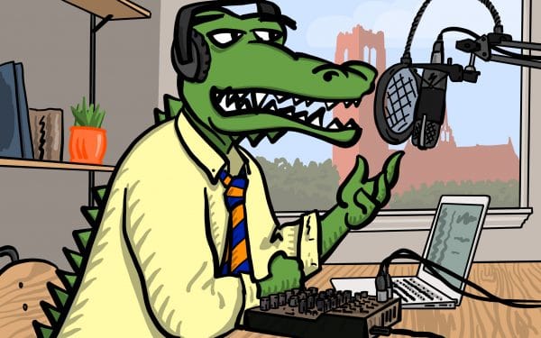 Albert Gator wearing a yellow shirt and orange and blue tie speaking into a microphone and wearing headphones while recording a podcast. Century Tower is in a window behind him.
