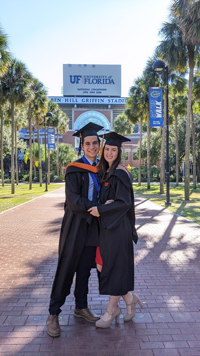 Sara Londono and her husband pose for a photo in front of Ben Hill Griffin Stadium. They wear graduation caps and gowns. 