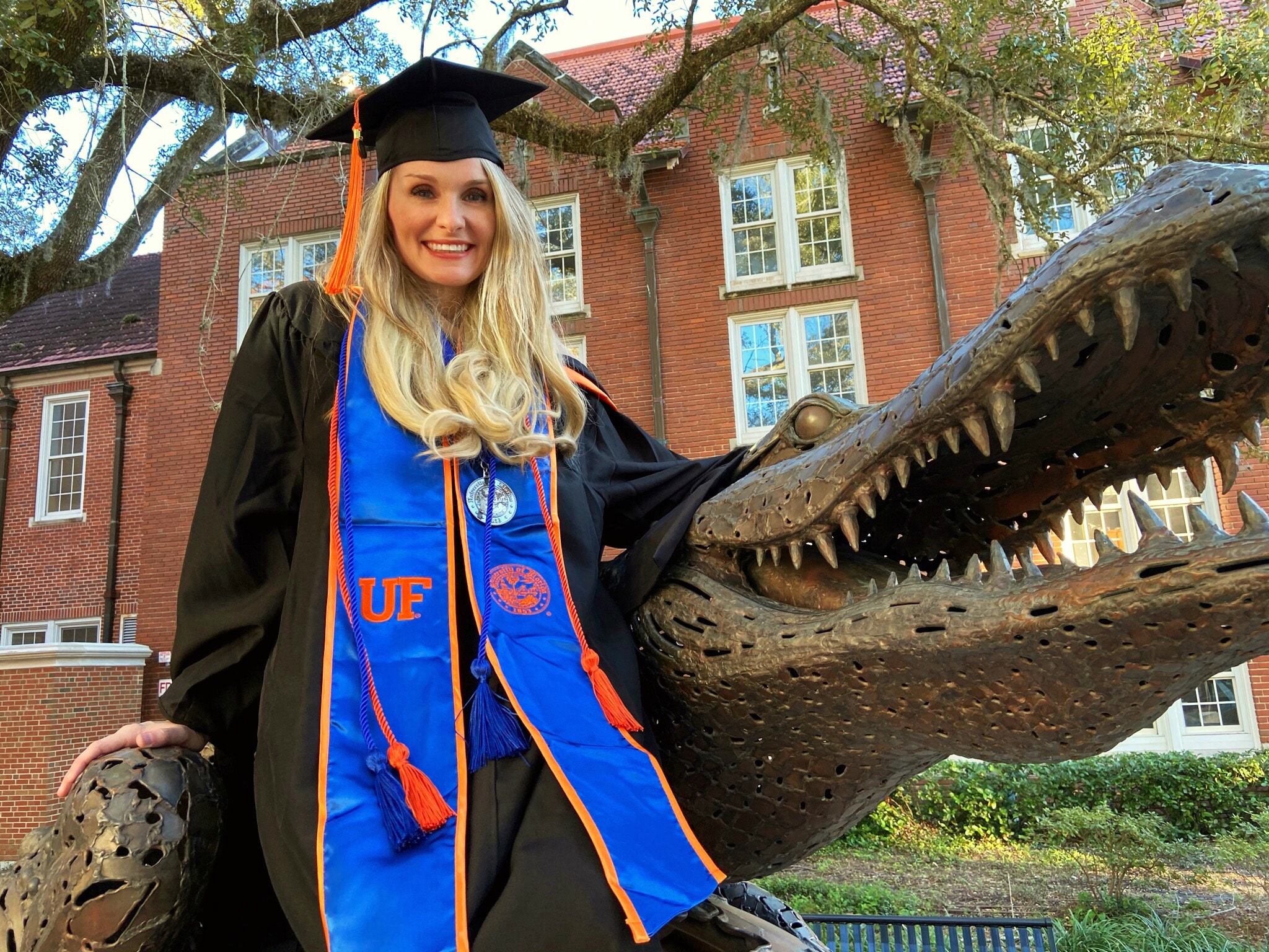 Katie Hudon wears a cap and gown with blue stole while sitting next to a large Gator statue.