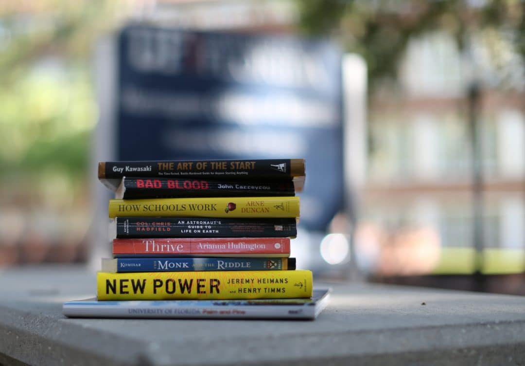 Stack of books on a picnic table in front of the Warrington College of Business sign.