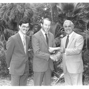 Two men pose for a photo with Robert Lanzillotti. One shakes Lanzillotti's hand while handing him a check.