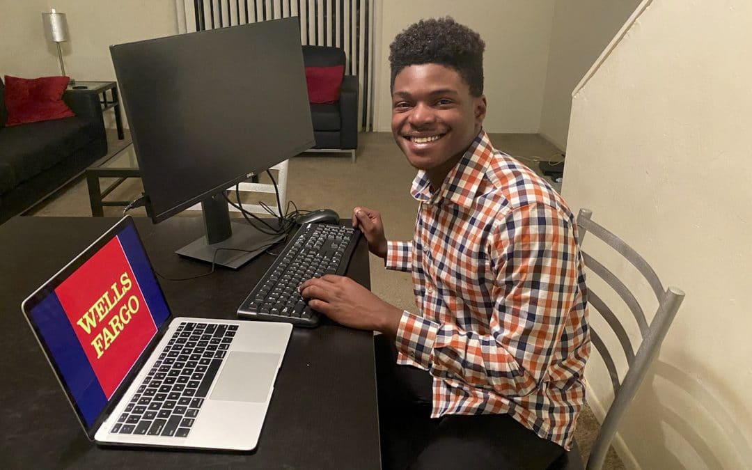 Dwayne Thelwell poses for a photo at his home work set up with a laptop and computer monitor with the Wells Fargo logo