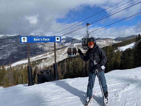 Benjamin Shu poses for a photo while skiing on a mountain next to a ski run trail sign that reads "Ben's Face"