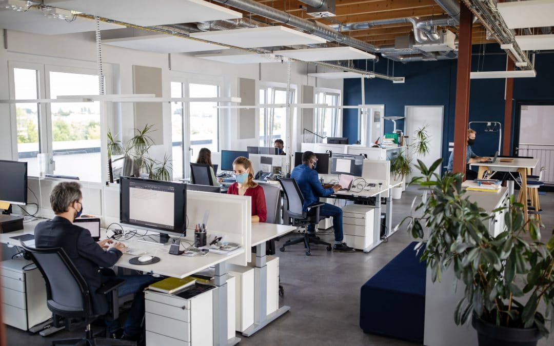 A large open office space with rows of desks. People sit at the desks working while wearing face masks.