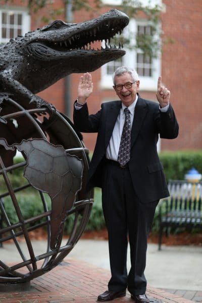 Wayne Archer holds up a '5' and '1' with his fingers while posting next to a large statue of a gator laying on top of a globe. 