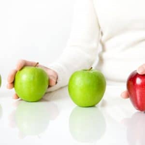 Four apples - three green and one red - sit in line with a person's hands on one green and one red apple as if trying to make a selection between the two.