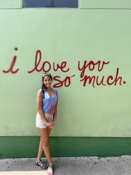 Tara Routie stands in front of a wall with the words 'I love you so much' spray painted on top.