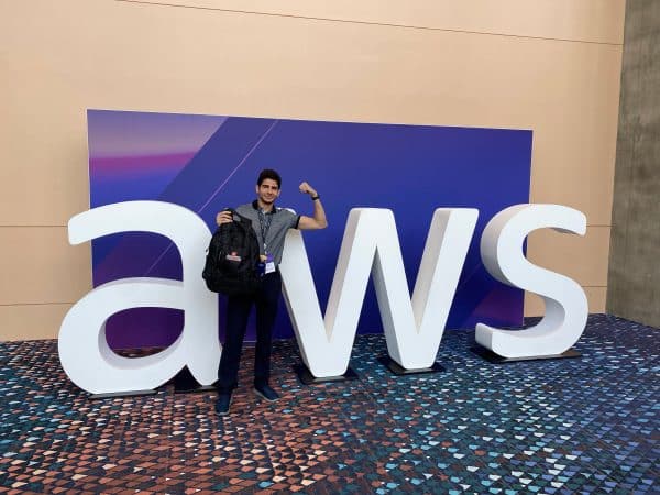 Alexia Munguia poses in front of a large AWS logo while holding up his MI-ISOM backpack