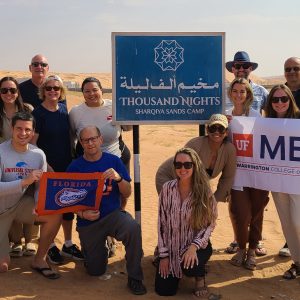 A group of about 15 people, including students and alumni, pose for a photo in the Omani desert while holding a UF MBA and University of Florida flag.