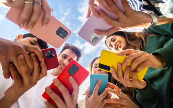 Young group of people using mobile phone device standing together in circle outdoors. Millennial friends addicted to social media app, betting or playing video game on platform online.