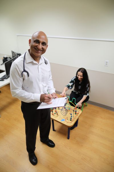 Sanjeev Tuli stands holding a clipboard while Sonal Tuli sits at a child's toy.