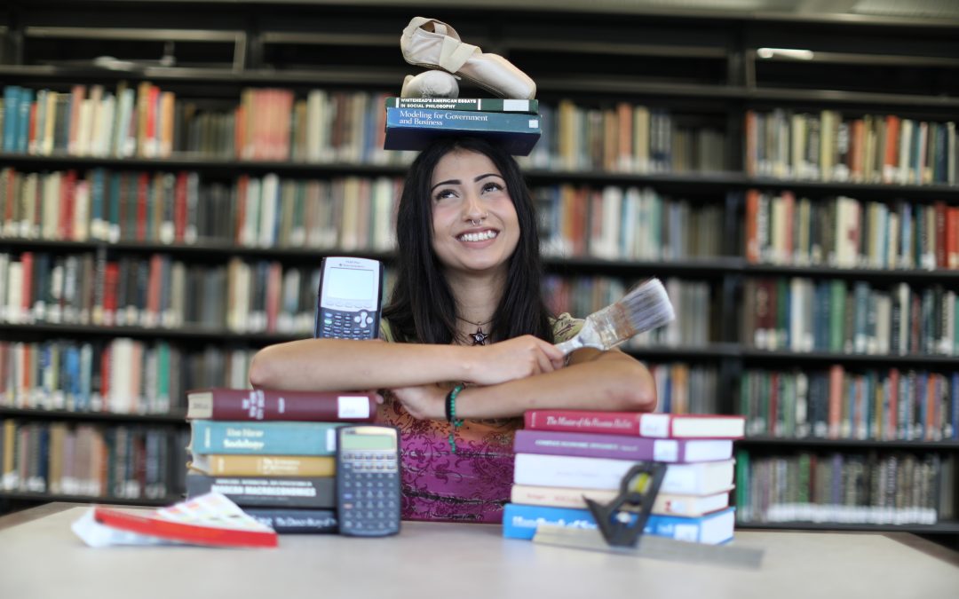 A student holds various objects while balancing books and ballet shoes on her head.
