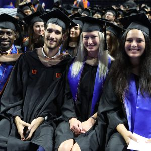 Four students, two male and two female, sit together in graduation regalia during a graduation ceremony.