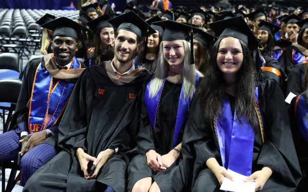 Four students, two male and two female, sit together in graduation regalia during a graduation ceremony.