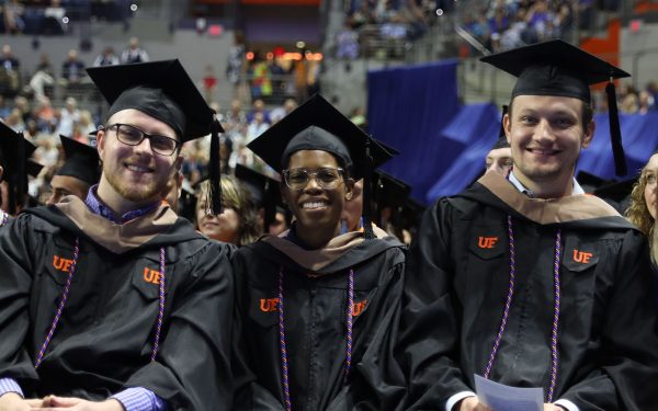 Three students wearing graduation regalia sit in a row during a graduation ceremony.