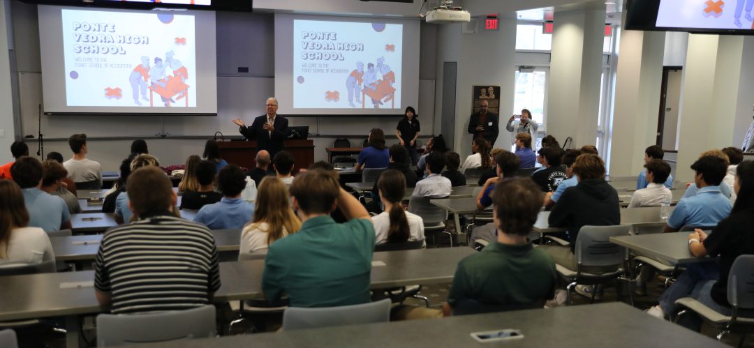 Students sit in a large auditorium classroom listening to a presentation.