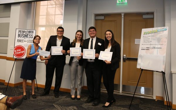 Five people holding certificates, standing indoors. A woman to the left gestures enthusiastically near a poster about business.