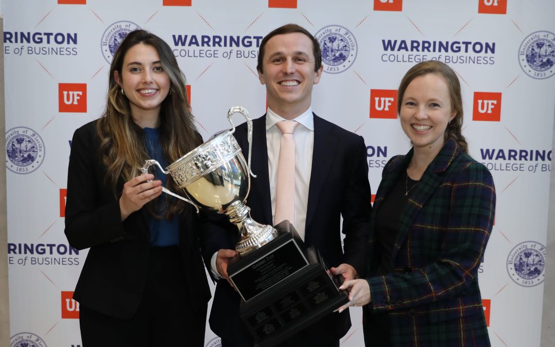 Three students wearing business suits hold a large trophy cup in front of a step and repeat with the UF Warrington College of Business logo