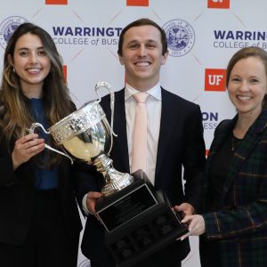 Three students wearing business suits hold a large trophy cup in front of a step and repeat with the UF Warrington College of Business logo