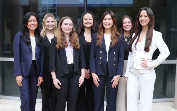 A group of seven students in business attire smiling in front of Hough Hall.