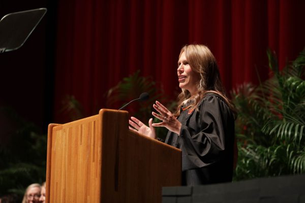 Jessica Fleisher wears a graduation cap and gown while speaking at a podium.