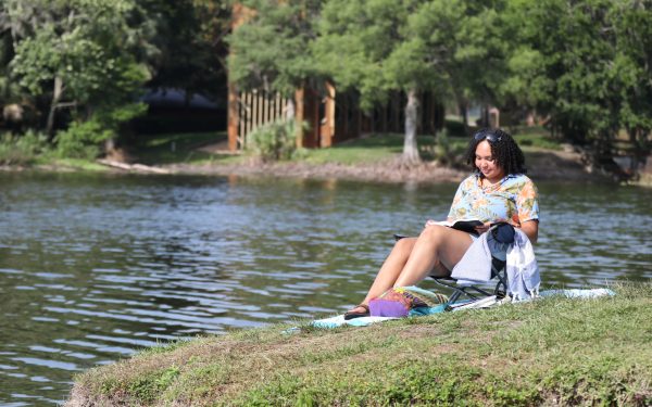 A young woman sits by a lake reading a book on a sunny day.
