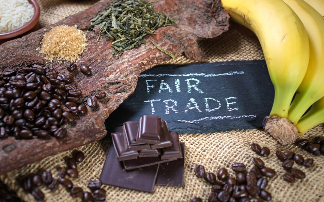 Assorted fair trade products including coffee beans, bananas, and chocolate displayed around a chalkboard sign that reads 'FAIR TRADE' on a burlap surface